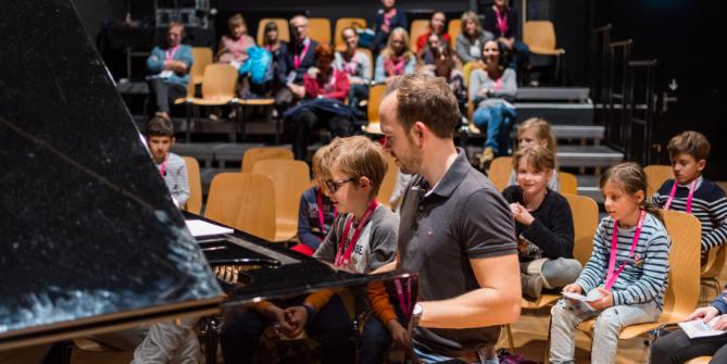 Kind mit Lehrer am Klavier, andere Kinder schauen zu