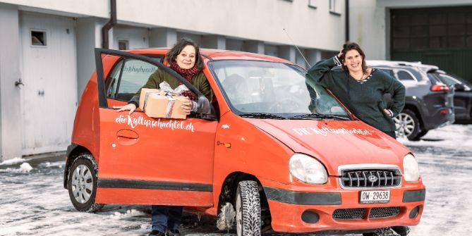 Les deux fondatrices de DIE Kulturschachtel se tiennent de chaque côté de leur voiture personnalisée, l’une tient un colis dans la main