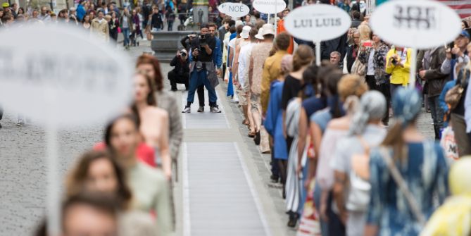 Une équipe de photographes se tient entre deux rangées de modèles qui défilent devant le public, sur une grande place en plein air.